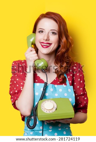 Smiling redhead girl in red polka dot dress and blue apron with green dial phone on yellow background.