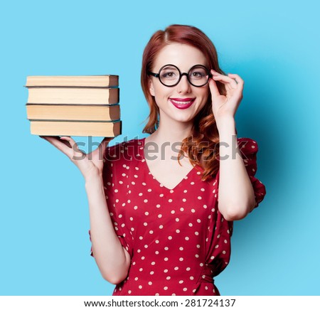 Young smiling redhead teacher in red polka dot dress with books on blue background.