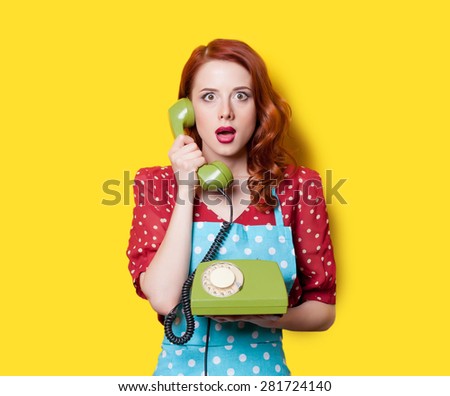 Surprised redhead girl in red polka dot dress and blue apron with green dial phone on yellow background.