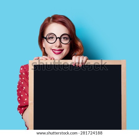 Young smiling redhead teacher in red polka dot dress with blackboard on blue background.