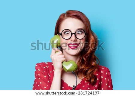 Smiling redhead girl in red polka dot dress with green dial phone on blue background.