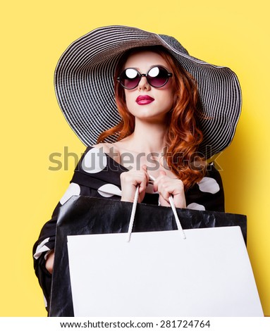 Redhead girl in black dress and hat with shopping bags on yellow background