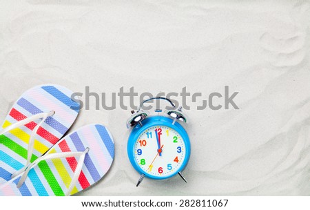 Colorful flip flops and classic alarm clock on white sand. Photo with high angle view.