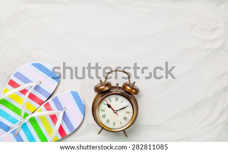 Colorful flip flops and classic alarm clock on white sand. Photo with high angle view.