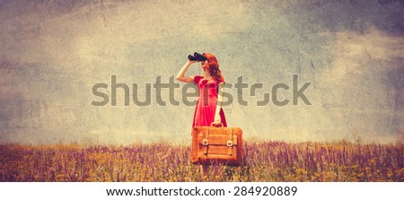 Portrait of a beautiful redhead girl in red dress with suitcase and binocular on the meadow. Photo in old color image style