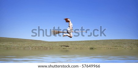 Young red-haired witch on broom flying over green grass field and water