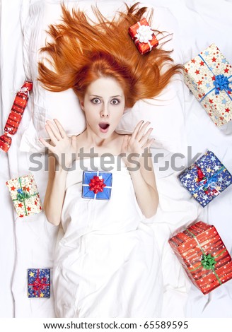 Surprised red-haired girl in bed with Christmas gifts. Studio shot.