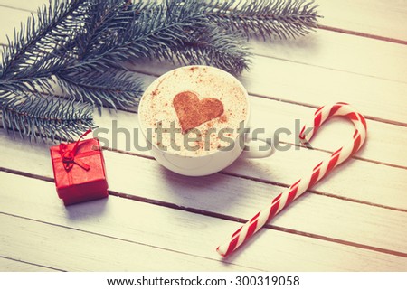 Cup of coffee with heart shape and christmas candy near red gift and brench on white wooden background.
