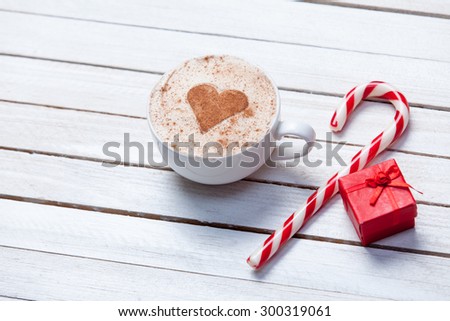 Cup of coffee with heart shape and christmas candy near red gift on white wooden background.