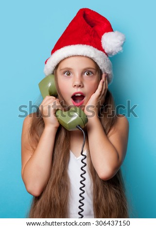 Young surprised girl in Santas hat calling by phone on blue background.