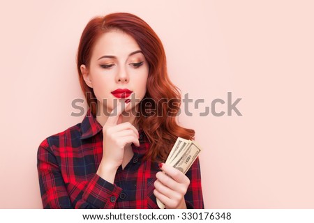 Surprised redhead girl in red tartan dress with money on pink background.