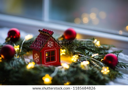 Red wooden toy house surrounded with fir-tree wreath decorated with warm garland lights and little Christmas balls near window. New Year festive glowing decoration. Concept of cozy home in winter.