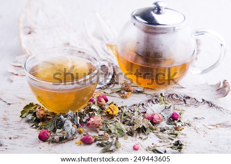 Transparent hot cup of herbal tea with teapot and dry roses on the tree bark