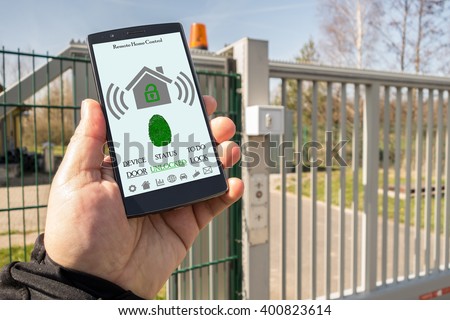 hand holding black mobile smart phone with smart home application on the screen. Blurred gate on the background. remote home control app on a smartphone. All screen graphics are made up.