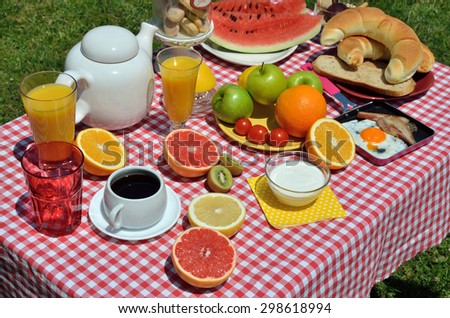 Breakfast or brunch with coffee, bacon and eggs, croissants, bread, cookies, cereals, Greek yogurt, orange juice, water-melon, orange, grapefruits and apples on red-white tablecloth served outdoor