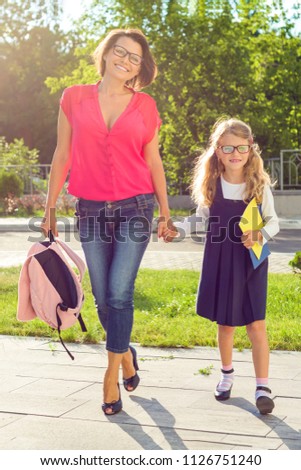Outdoor portrait of mother and child going to school. Girl 6, 7 years old with glasses, school uniform and happy mom holding hands, back to school