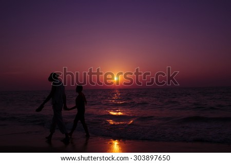 Loving couple walks hand-in-hand on the beach with a beautiful sunset in front of them