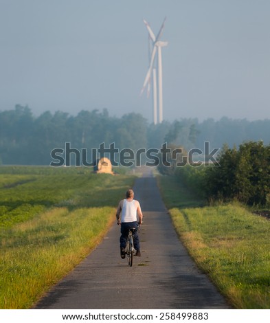 Old country road in soft focus and cyclist going forward. On background wind turbine - renewable energy source in Germany