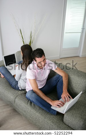 Portrait of a man and a woman sat on a sofa back to back in front of a laptop computer