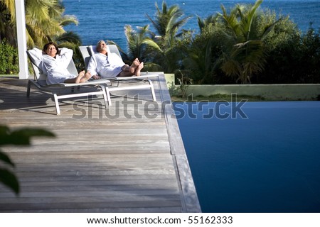 Senior couple in bathrobe lying on deck chairs next to a swimming pool