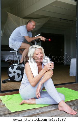 Elderly woman resting with a glass of water near a man doing bike