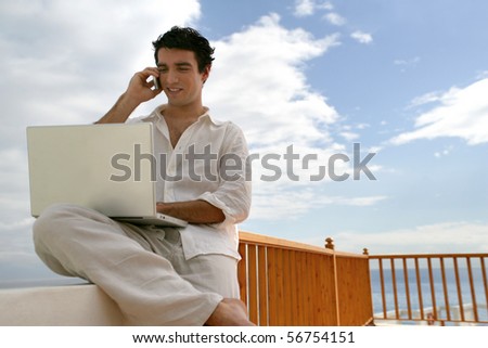 Young man phoning in front of a laptop computer on a terrace