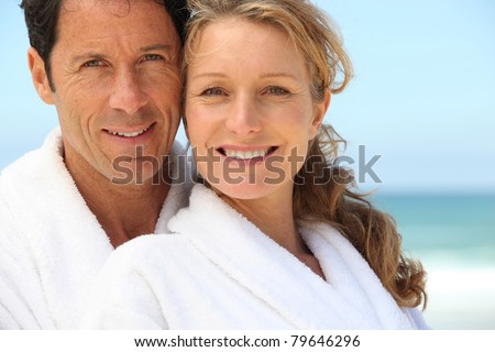 A man and a woman wearing dressing gowns and smiling at us on a beach.