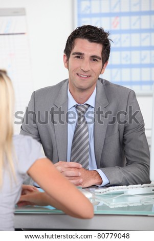 Man in suit sitting across a desk from a young woman with a schedule in the background