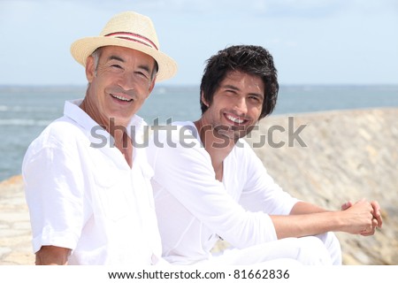65 years old man and a 30 years old man sitting on the sand with background of sea