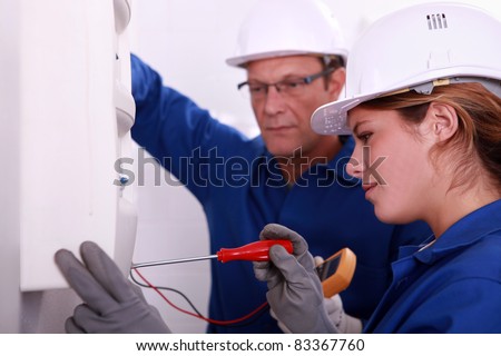 a young woman working on an electricity meter accompanied with a 45 years old man, both are wearing blue jumpsuites