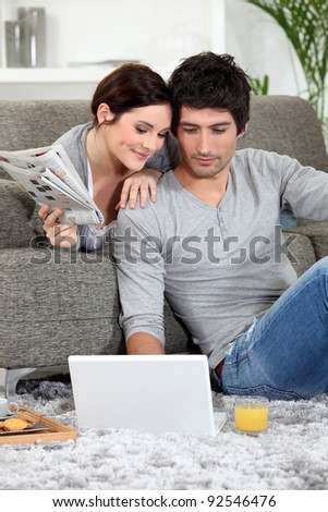 Couple sat on couch with newspaper and laptop