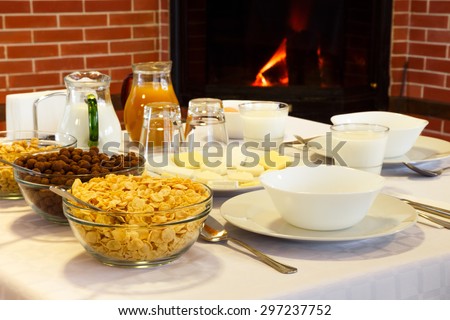 Hotel breakfast with corn flakes, milk, cheese, yellow cheese and orange juice on a hotel table near the fireplace comfort.