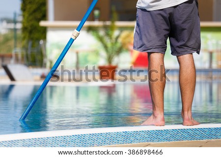 Man cleaning a swimming pool in summer with a brush or net on a blue pole standing barefoot on the tiles at the edge, low angle view of his lower body.