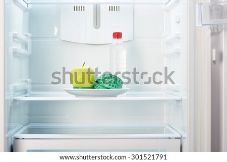 Green apple with measuring tape on white plate and a bottle of water in open empty refrigerator. Weight loss diet concept.
