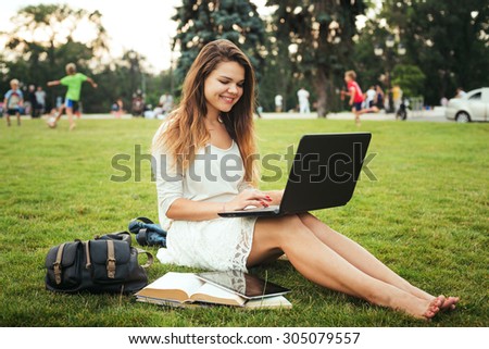 young caucasian female student sitting on a grass in campus area near the university, holding a laptop on her knees. Books, backpack and tablet is around. Student study in campus, education concept