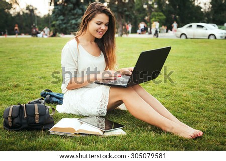 young caucasian female student sitting on a grass in campus area near the university, holding a laptop on her knees. Books, backpack and tablet is around. Student study in campus, education concept