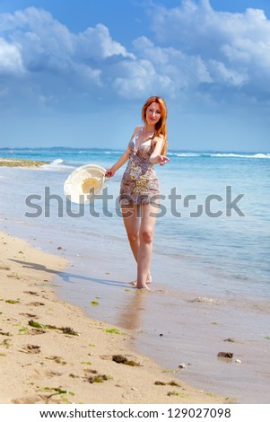 Young beautiful woman goes on ocean coast with straw hat in hands