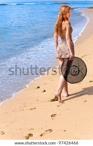 Young graceful woman goes on coast of ocean with straw hat in hands