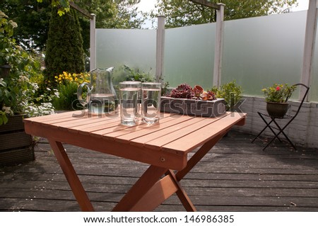 Wooden terrace table with a carafe and three glasses filled with water. Garden plants at the background