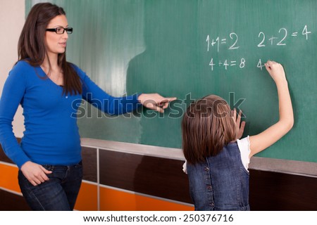Little girl in primary school writing mathematics task on chalkboard while teacher looking at her