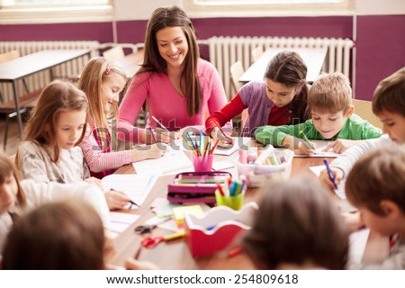 Pupil boys and girls sitting together around the table in classroom and drawing. With them is their young and beautiful teacher. She teaches children and smiling