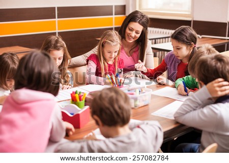 Children boys and girls sitting together around the table in classroom and drawing. With them is their young and beautiful teacher.