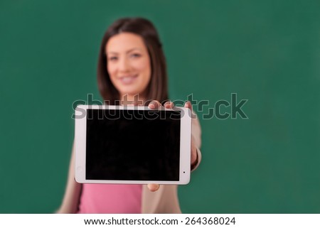 Young woman standing in front of empty chalkboard and holding a digital tablet and showing it. She is looking a the camera and smiling
