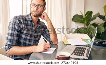 Young writer wearing a check shirt and glasses looking out at the view from his home office\'s window, taking notes every now and then from his notebook and a black coffee