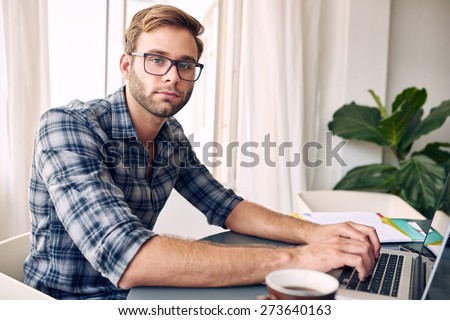 Young student wearing a check shirt and glasses looks into camera while typing on his new notebook, with a cup of coffee on the ready and files on his desk