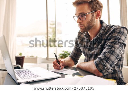 Student busy taking notes down from his new laptop computer while browsing the internet with his morning coffee