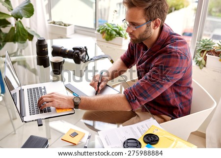 Young photographer retouching on his new laptop with his stylus and tablet on his glass desk, in his home studio