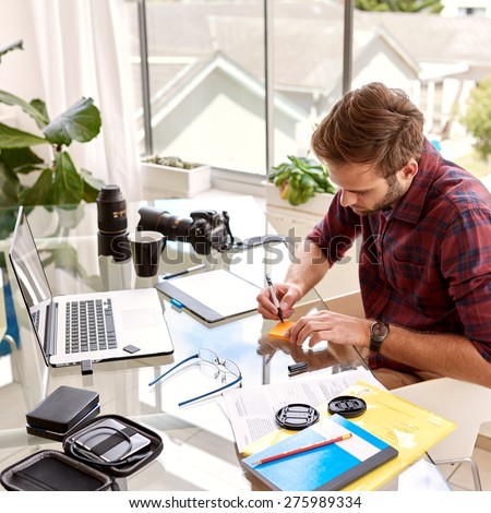 Young businessman busy taking notes on a note pad, sitting at his glass desk in his home office with his morning coffee