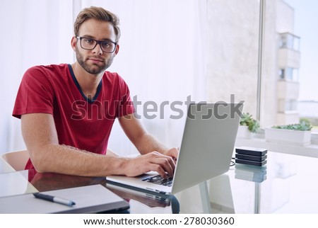 young photographer looking at the camera with a blank expression, while he works on his laptop computer with his red shirt and trimmed beard