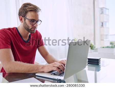 young student busy typing from his home studio on his new laptop computer while sitting at his glass desk with is red shirt and trimmed beard while wearing glasses